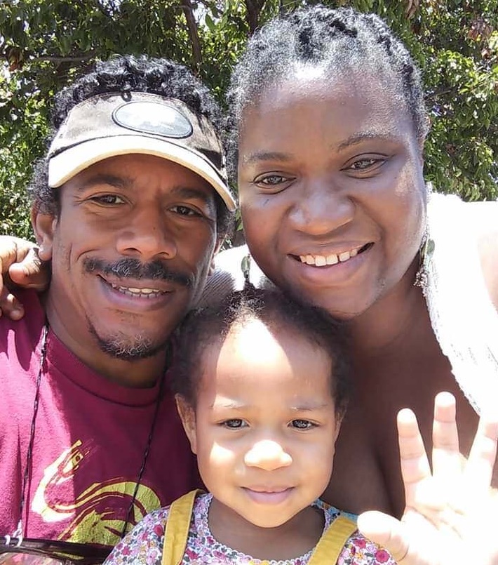 An African American family (man, woman, and toddler) smile at the camera in a selfie photo.