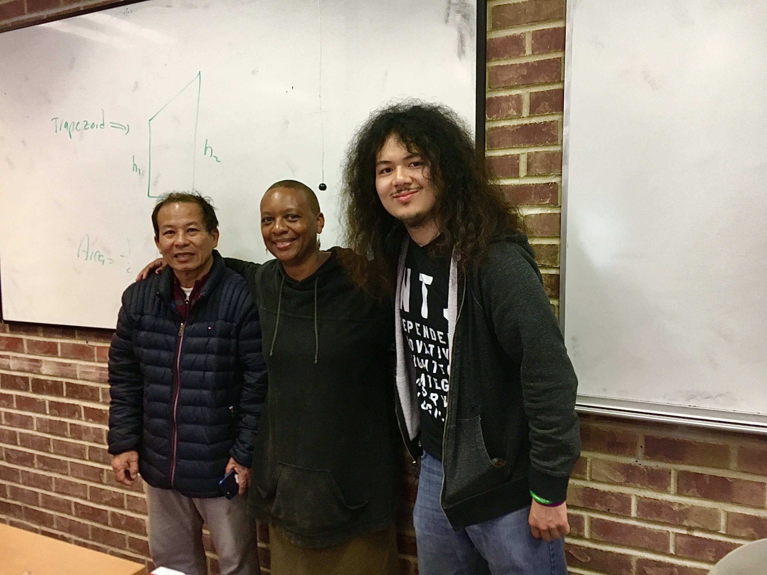 Three people line up in front of a whiteboard to take a photo: an elder Asian American man, an African American woman college instructor, and a young man with long hair.