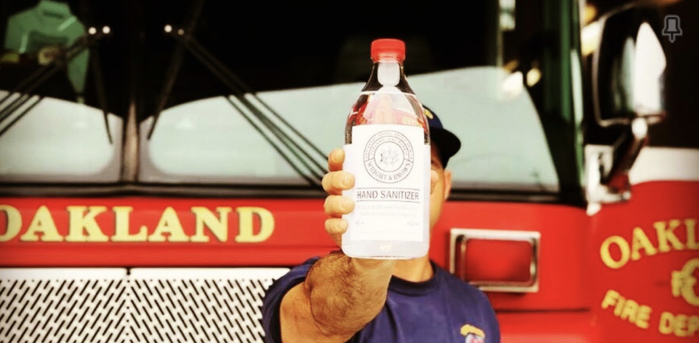 A firefighter is holding a glass container of hand sanitizer. In the back is a bright red firetruck that says "Oakland Fire Dept."