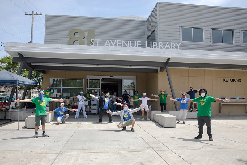 A diverse group of people wearing masks is standing in front of the 81st Ave library in East Oakland with their hands up and socially distanced.