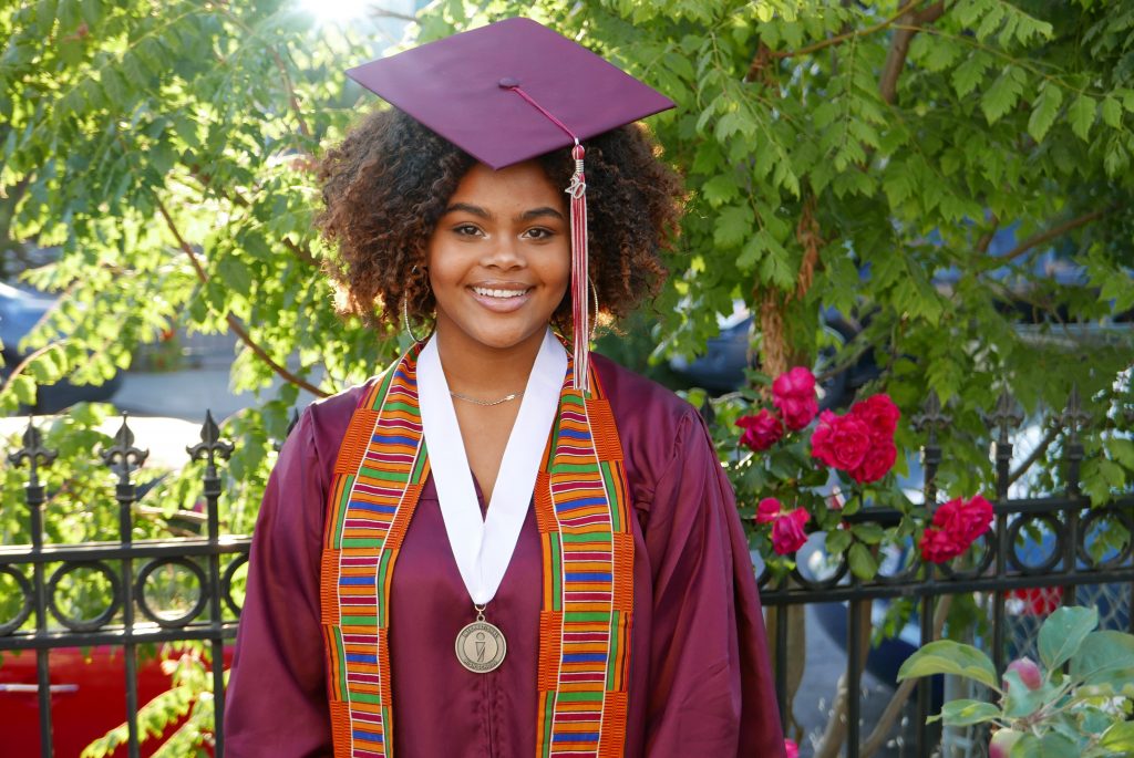 A smiling African American teenager wearing a maroon cap and gown smiles for the camera.
