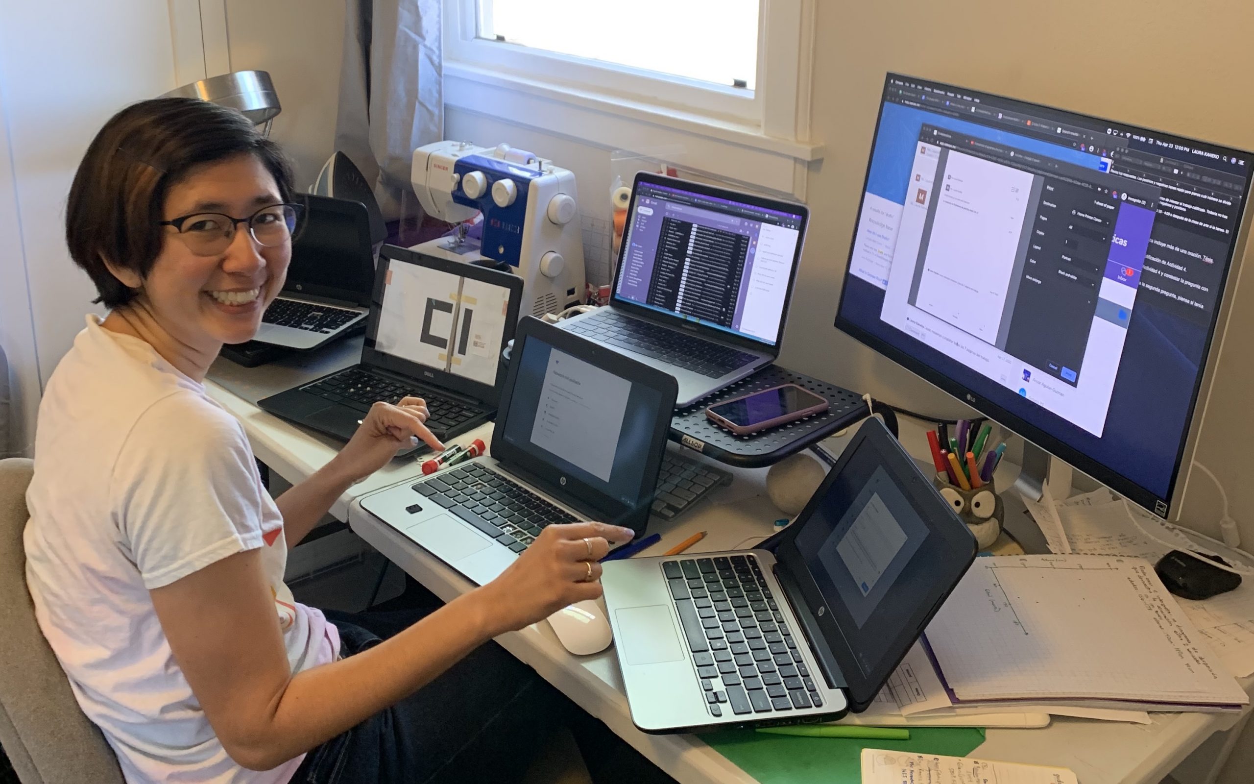 A smiling East Asian woman with a bob haircut and glasses is sitting at a large desk with several Chromebook laptops in front of her, plus a big monitor.