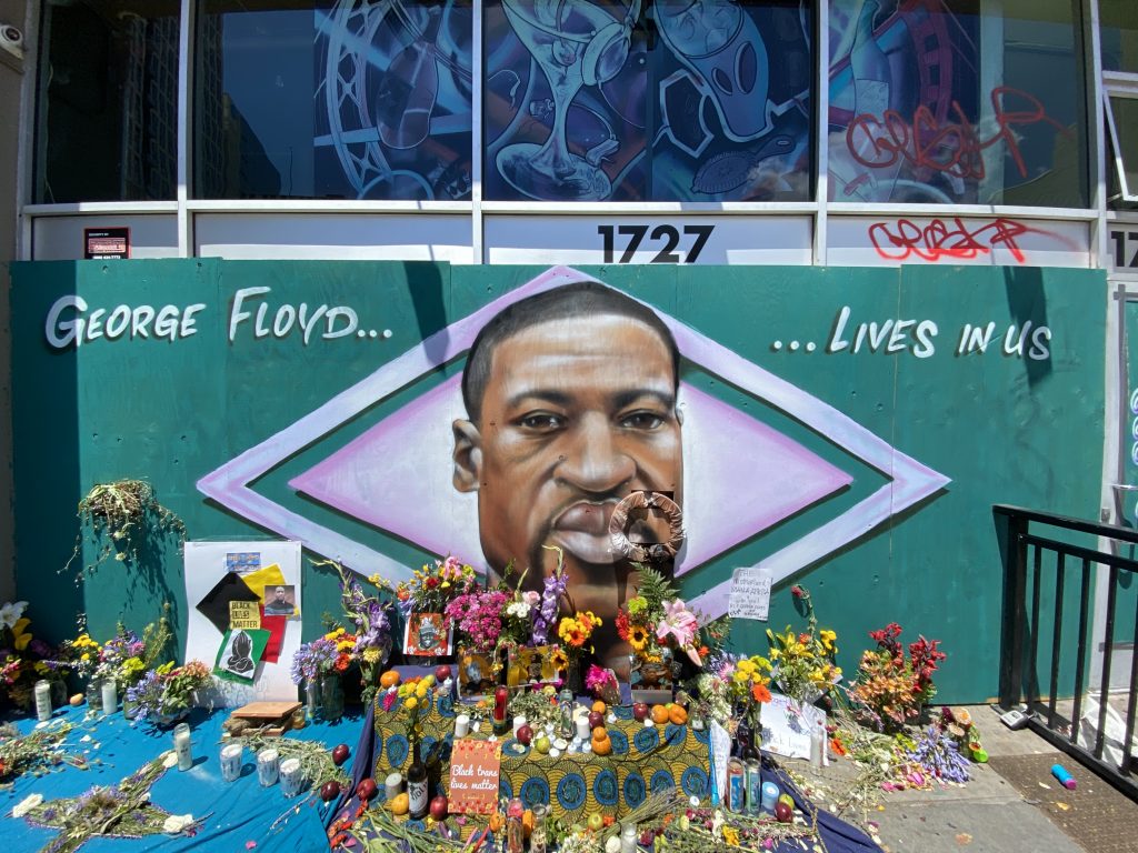 An image of an African American man at the center of a mural with a green background. Flowers are in front of it. It is an alter for the man who was killed by police.