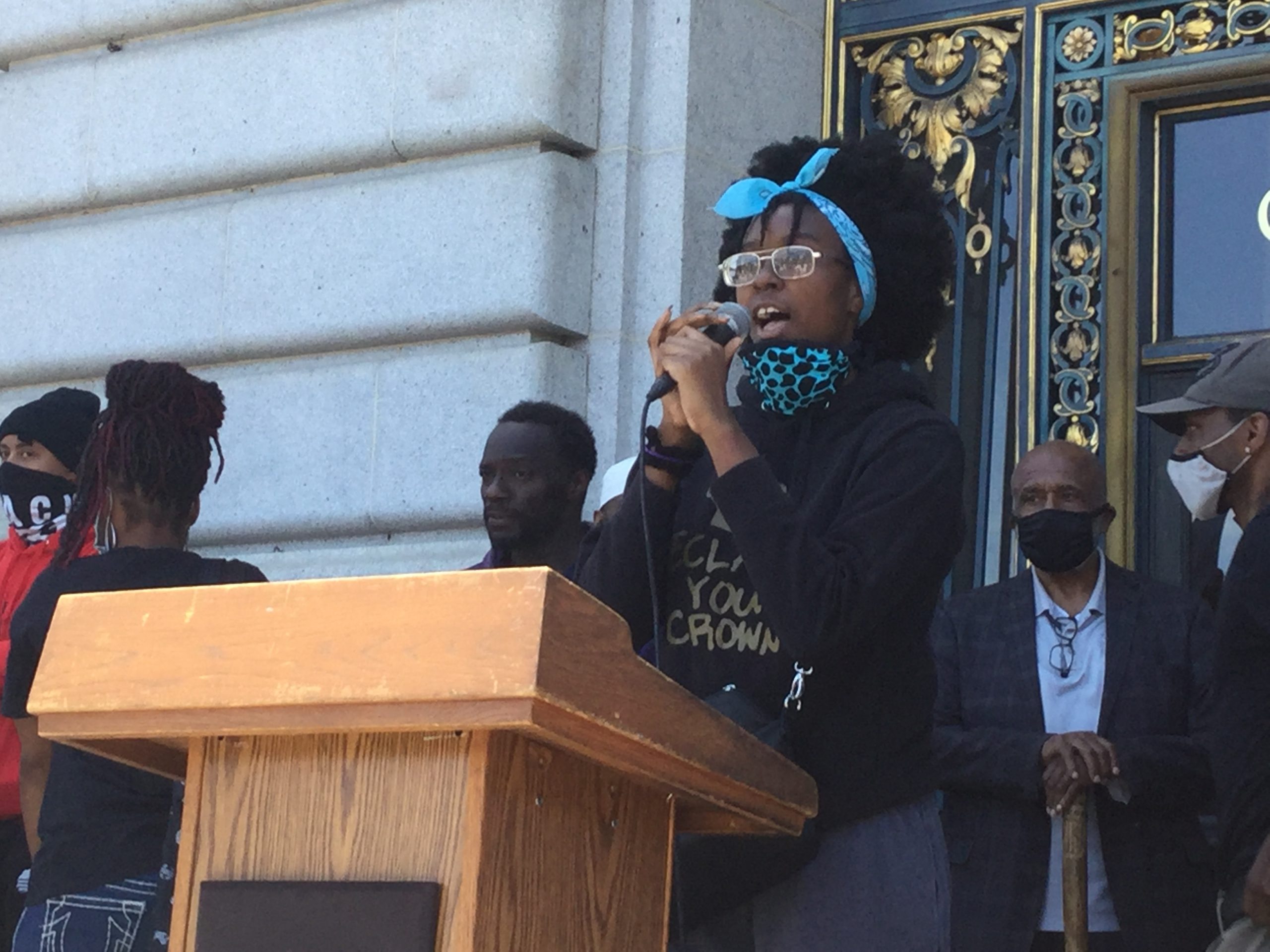 An African American teen girl, Alysia Reaves, speaks into a microphone behind a podium in front of SF City Hall.