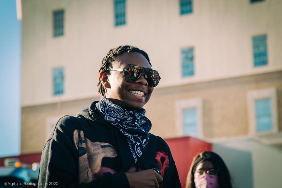 An image of a smiling African American youth who is wearing sunglasses and a bandana around his neck.