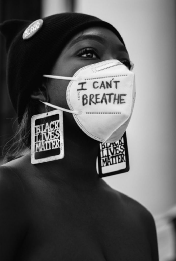 A young African American woman, Jacqueline Azah, looks into the distance. She is wearing a black beanie, a mask that says "I can't breathe," and earrings that say "Black Lives Matter."