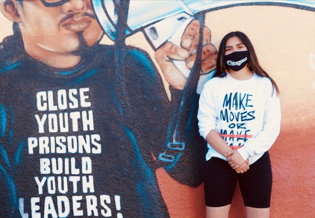 A Latina youth wearing a black masks stands in front of a mural that says "close youth prisons build youth leaders."