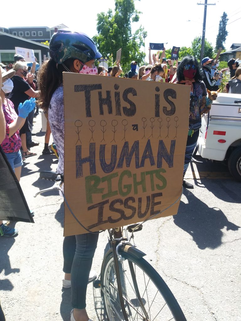 A cardboard sign is held up by someone new to a bike. The sign says "This is a human rights issue."