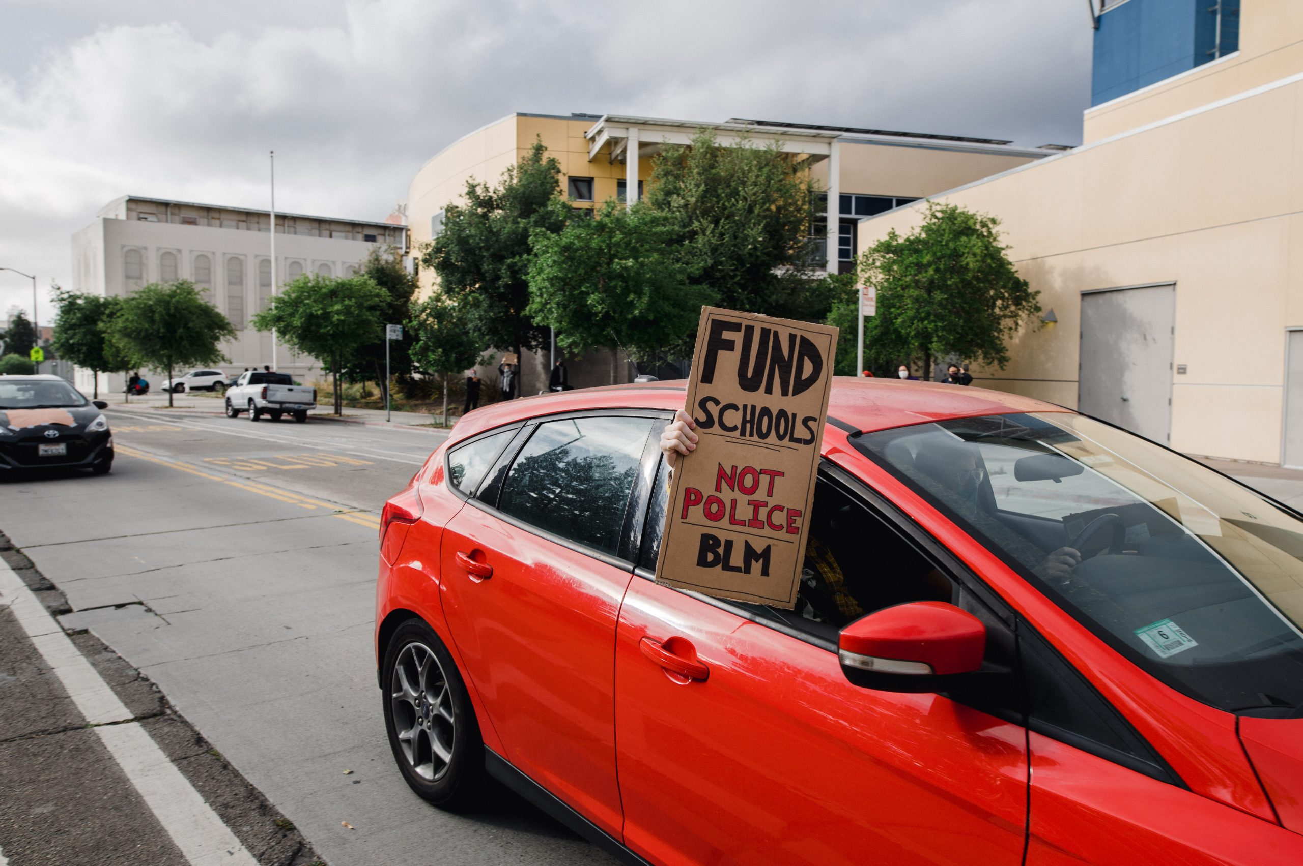 A red car has the window open. A hand is holding a homemade cardboard sign that says "Fund Schools Not Police BLM"