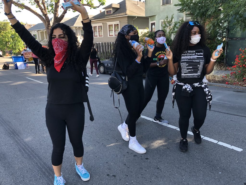 Several young women wearing black and with bandanas look happy and are passing out free water bottles on the street during a protest in Berkeley.