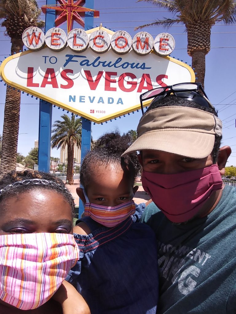 An image of a beautiful African American family (parents and young toddler girl) in front of a Las Vegas sign/marquee.