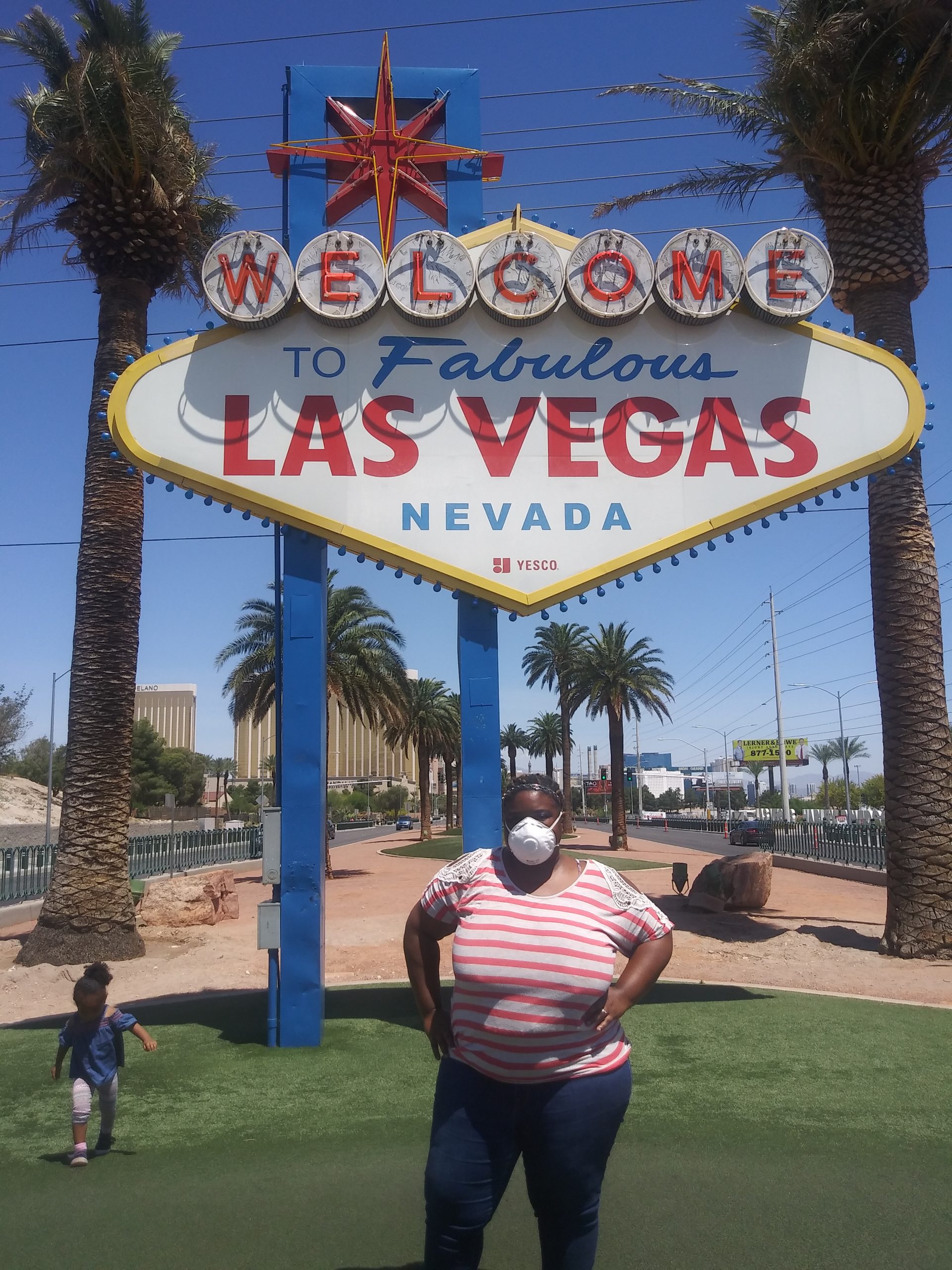 An African American woman stands in front of the Las Vegas sign, with a child running in the background
