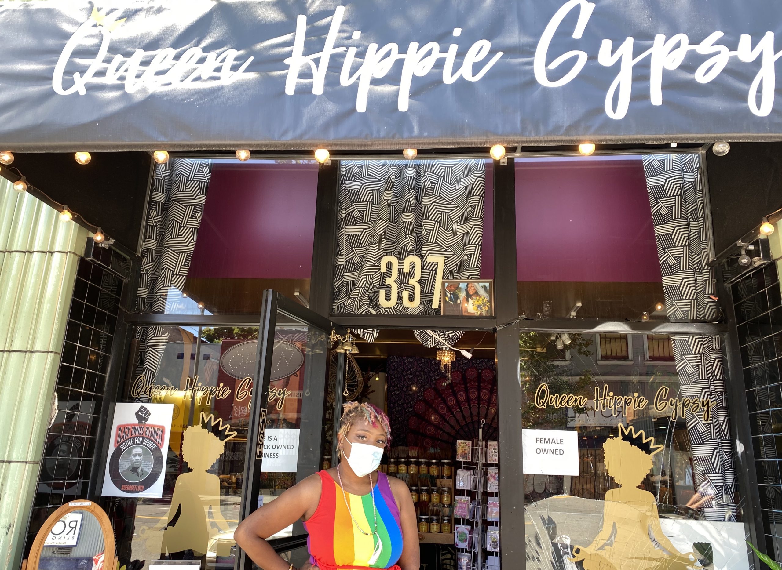 An African American woman wearing a rainbow colored dress stands in front of a storefront in Oakland.