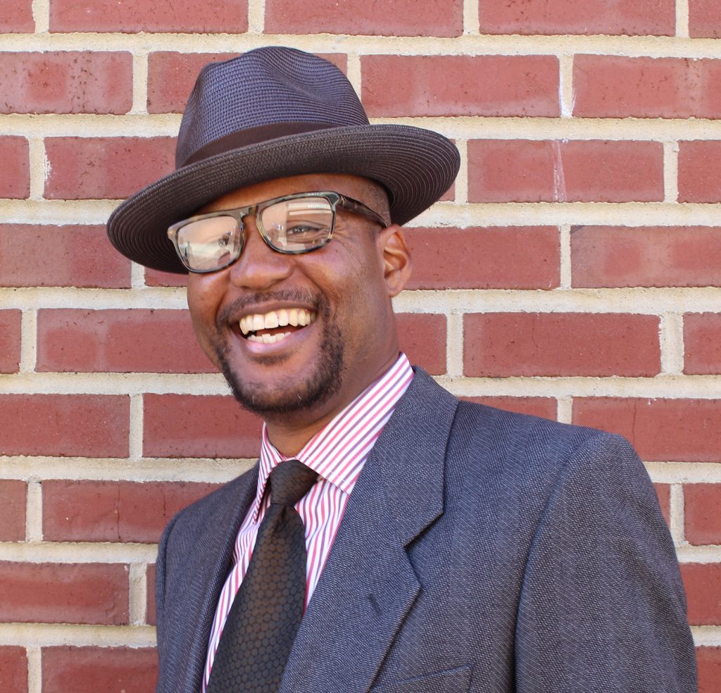 An African American man in glasses and a hat smiles in front of a brick background
