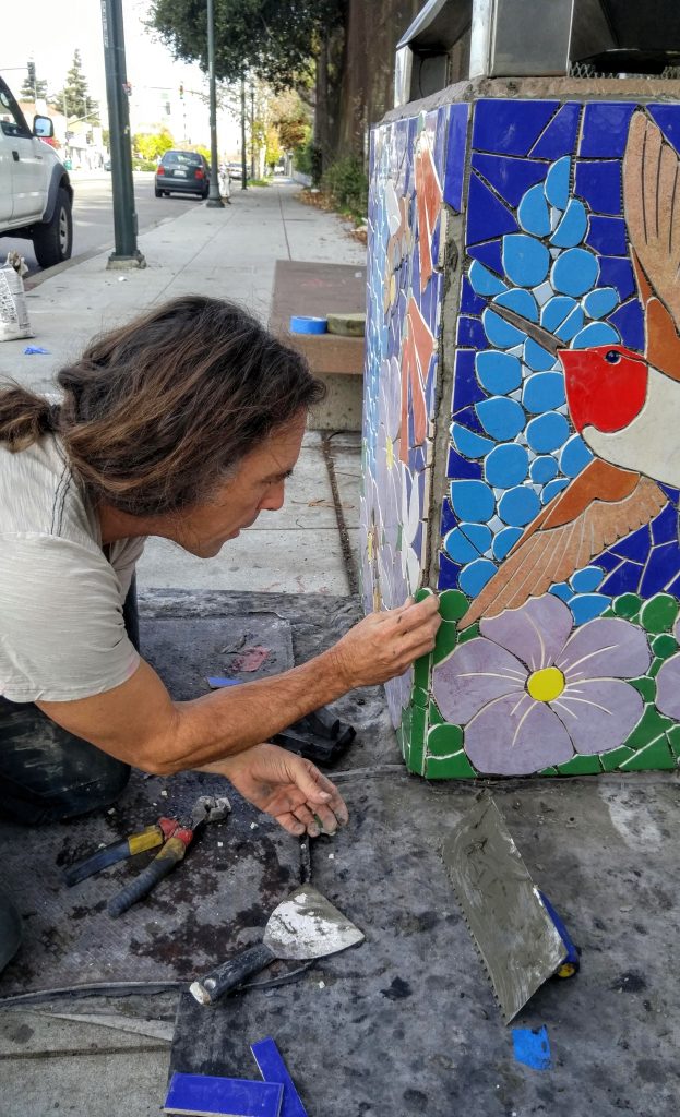 A male artist in a ponytail puts on mosaics on a trash can in Oakland.