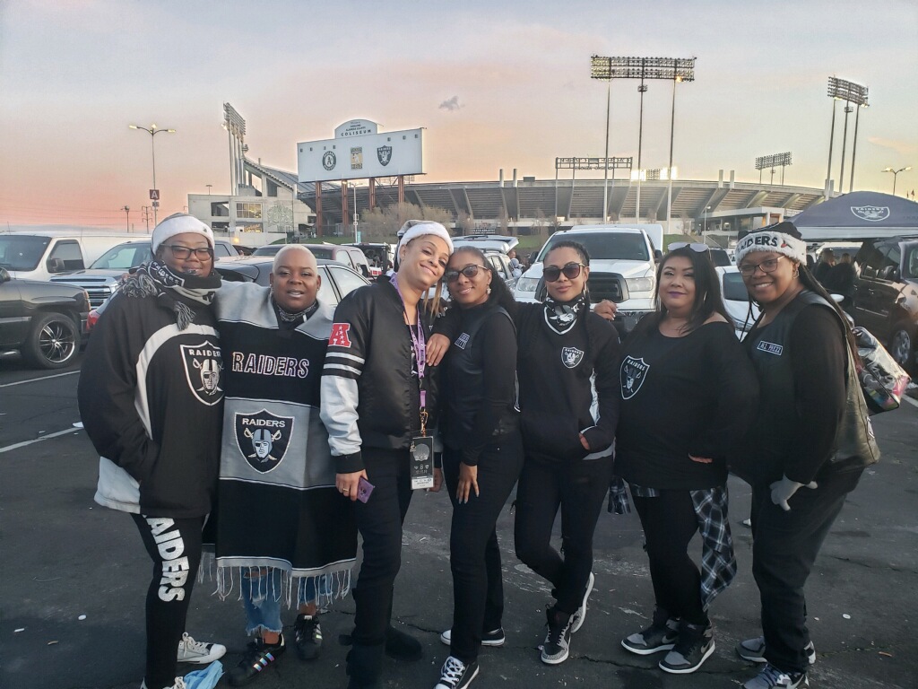 A group of women decked out in Oakland Raiders gear pose smiling in a photo in the parking lot.