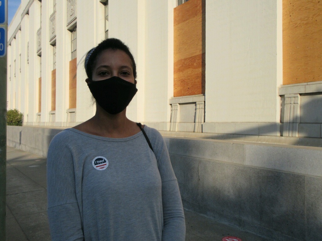 A young woman wearing a mask stand in front of a building wearing an "I voted" sticker