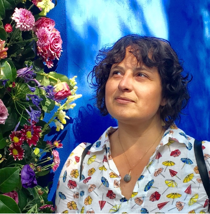 A woman with curly hair looks afar in front of a bright blue wall and flowers.