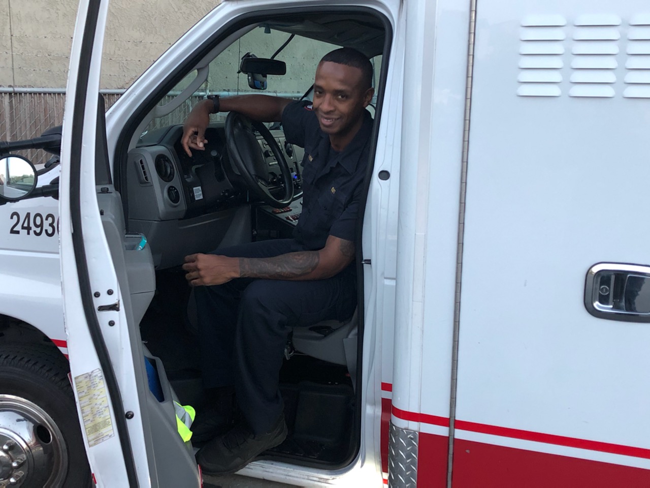 A young African American man sits in the driver's seat in an ambulance and smiles.