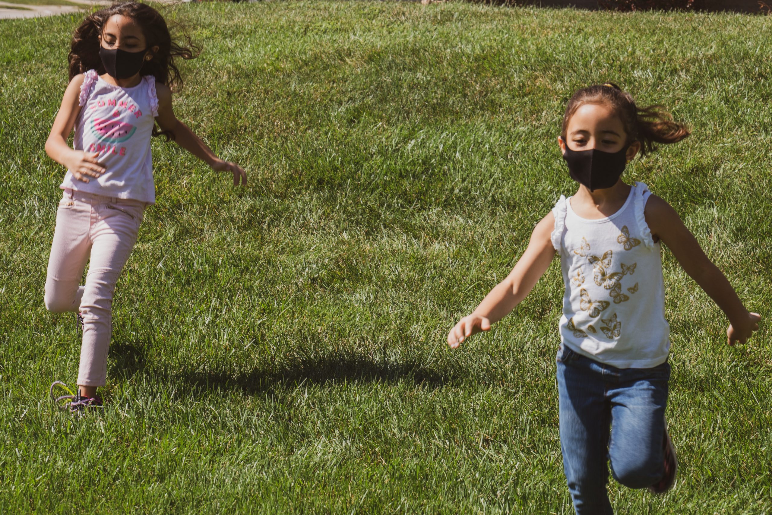 Two girls running on grass wearing black face masks.