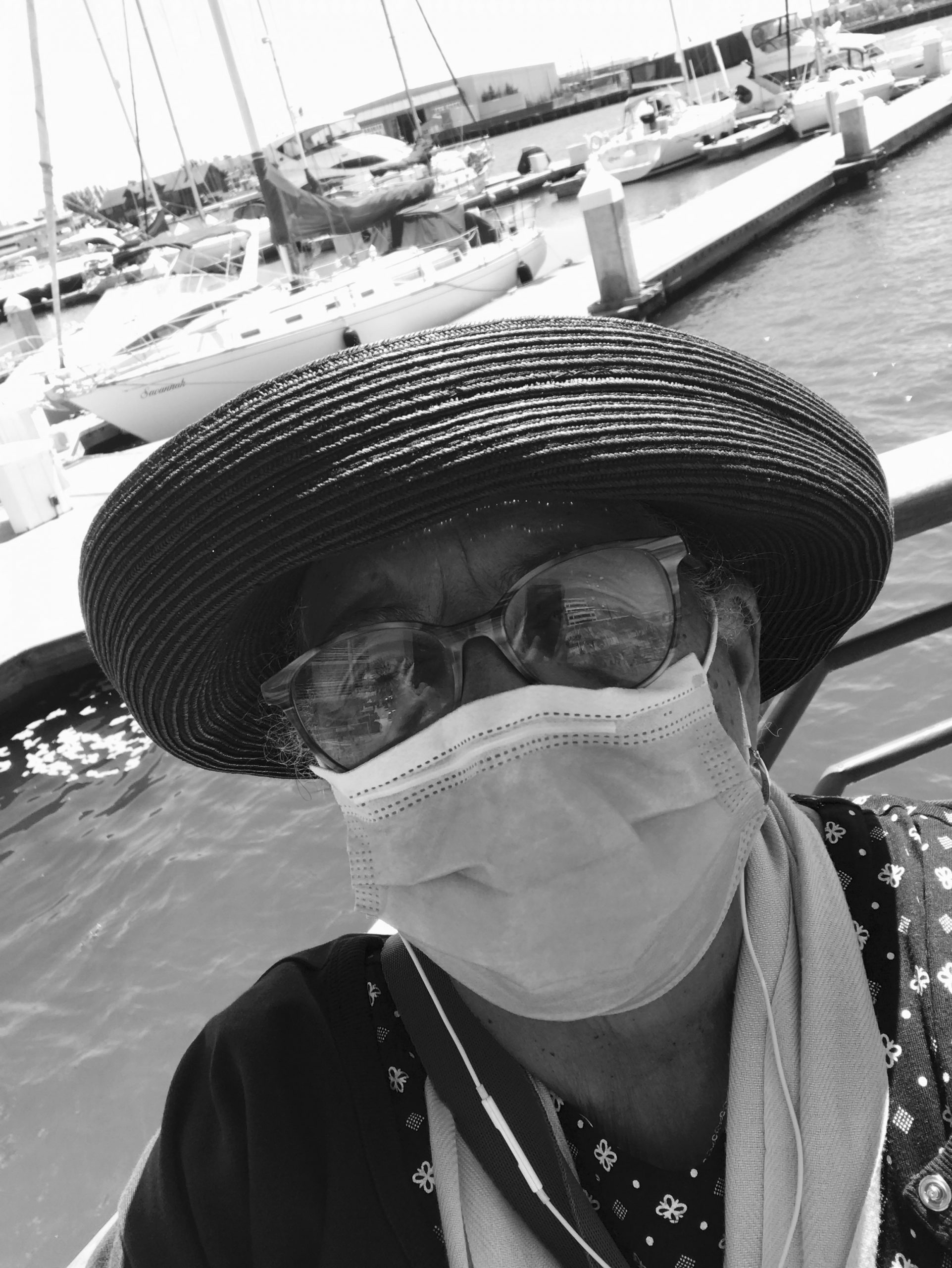 A black and white image of a woman wearing a hat, glasses, and a face mask standing outside by boat docks