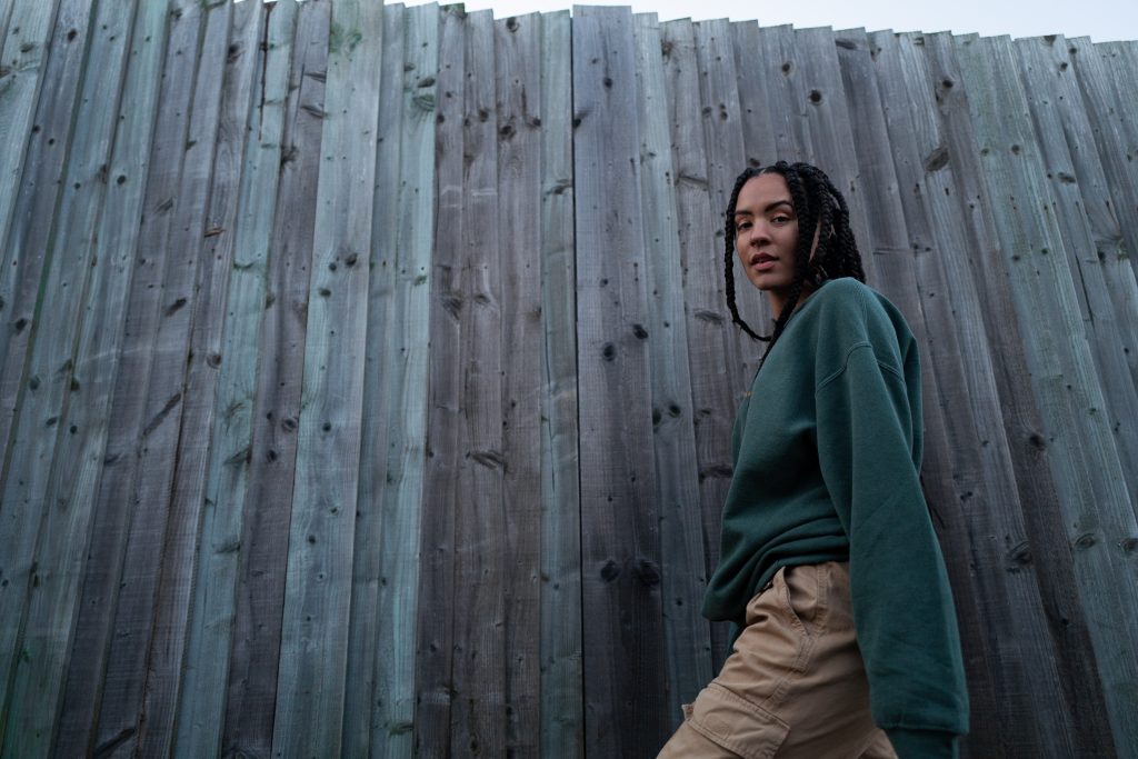 An image of an African American woman walking in front of a fence, with long braids and a green sweatshirt. She is smiling.