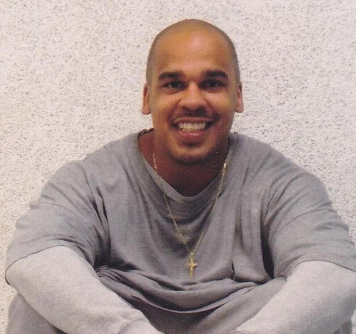 An African American man with no hair smiles at camera while sitting, wearing a grey inmate uniform.