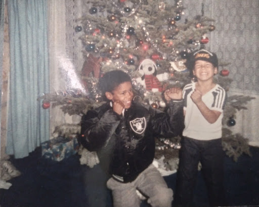 An image of two African American brothers play fighting in front of a Christmas tree and smiling, one wearing a tank top and the other a Raiders jacket.