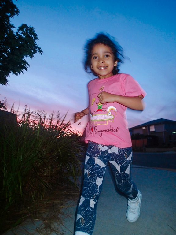 a sunset, a young girl wearing a pink shirt and blue leggings runs and smiles