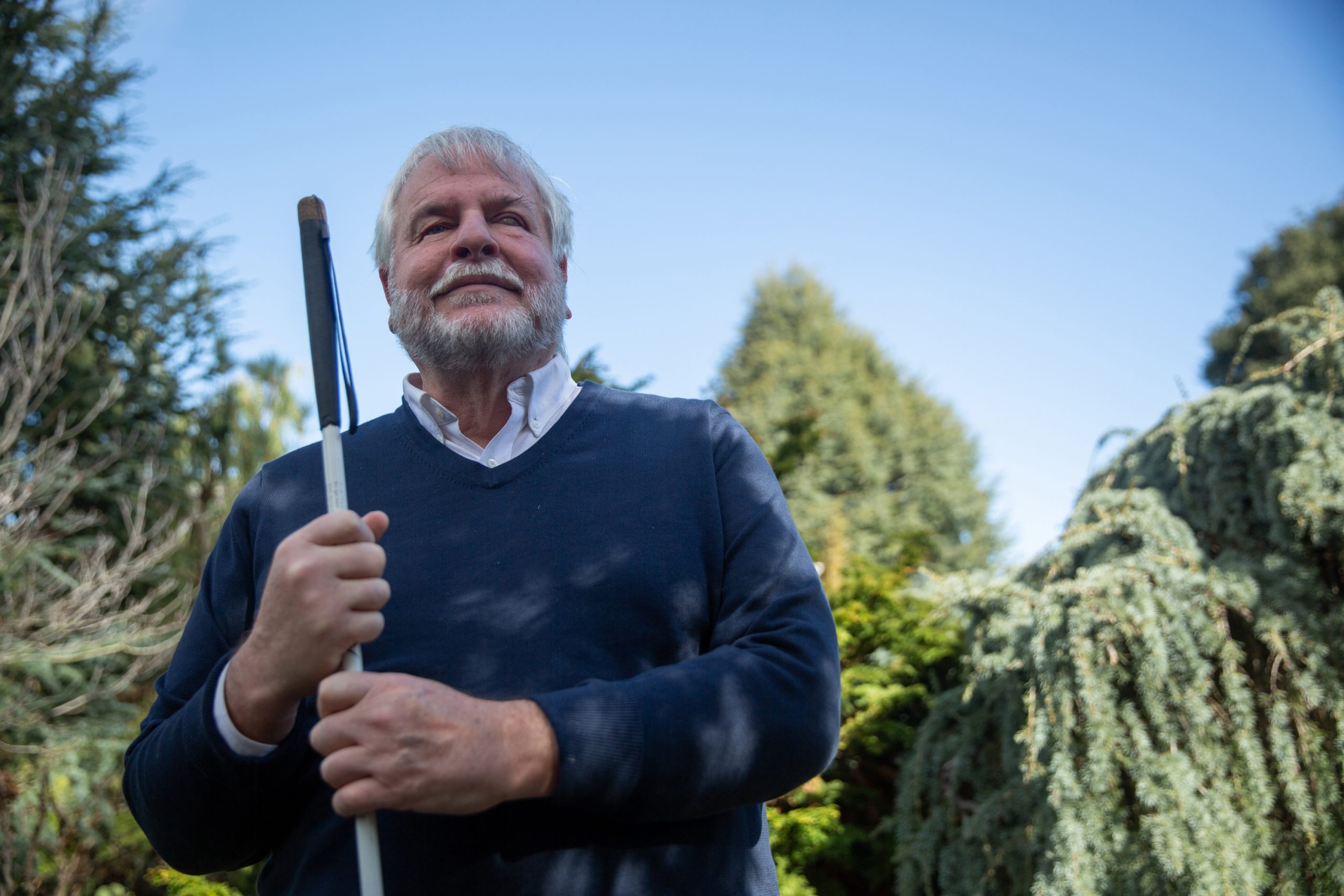 An image of a man who is blind with short grey hair, and green trees in the background.