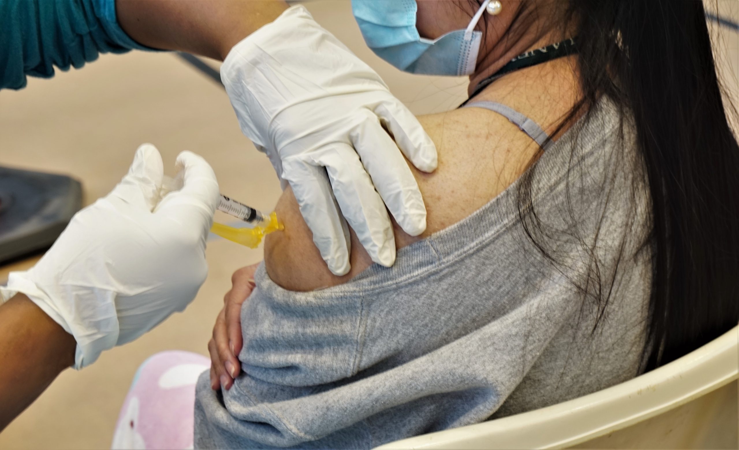 A closeup image of a young woman receiving a shot on her shoulder, with the person administering wearing latex gloves.