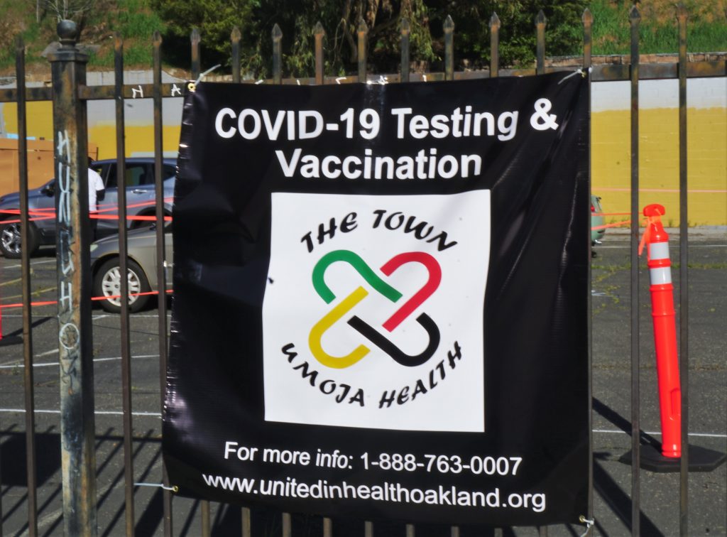 A square banner hands off of a fence that says "COVID-19 TESTING & Vaccination" Umoja Health.