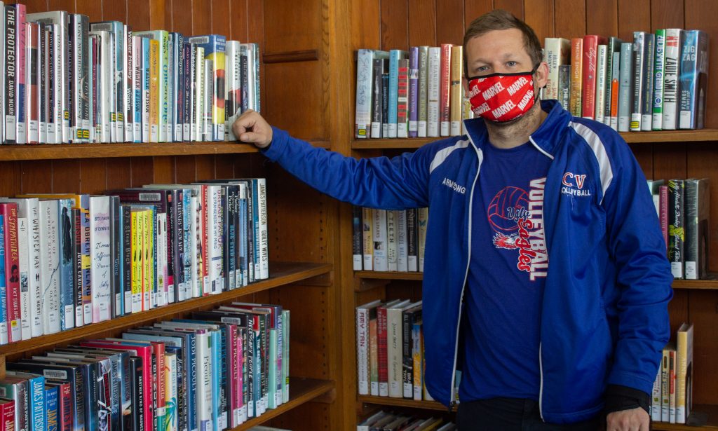 A man wearing blue jacket and red mask posed in front of a big bookshelf in the library.