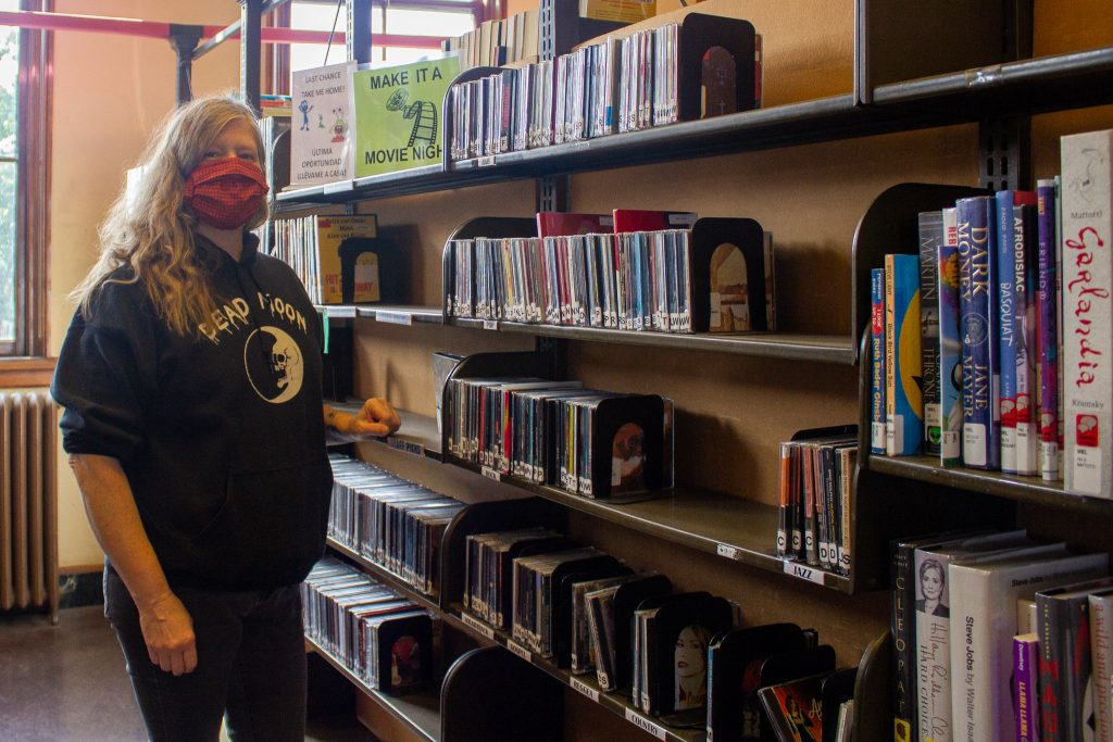 A library worker with wavy blonde hair wearing maroon colored mask and black hoodie stands by big bookshelves