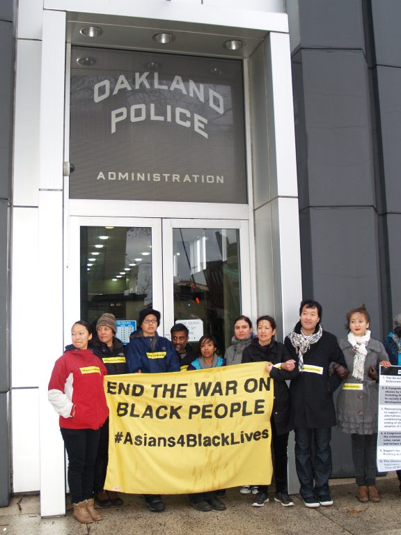 A group of Asian Americans hold a sign that says "End the War on Black People" and #Asians4BlackLives in front of the Oakland Police Department doors.
