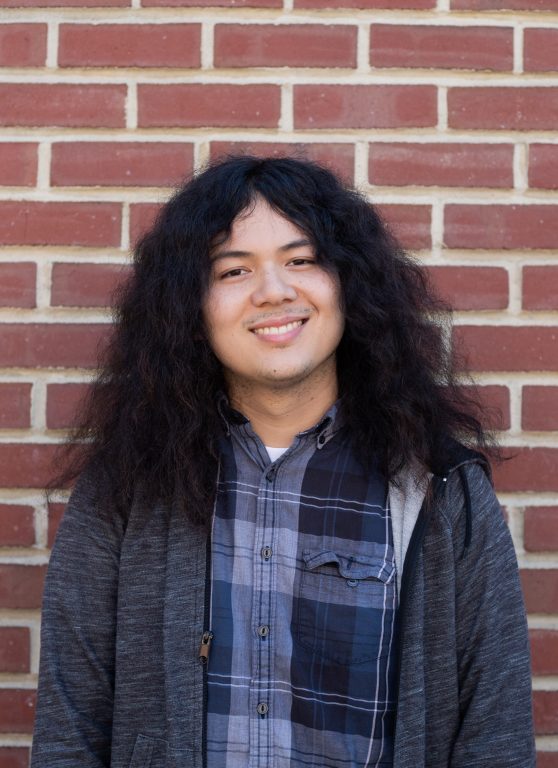 A young man with long, poofy hair stands in front of a brick wall and smiles for the camera.