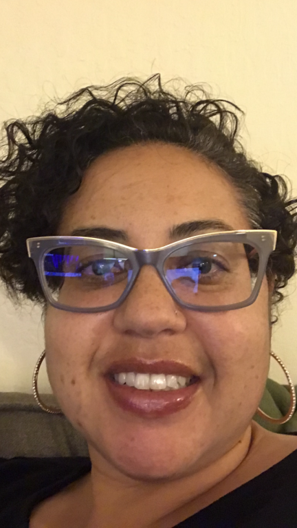 a woman with short brown curly hair, cat-eye glasses, and hoop hearings smiles at camera