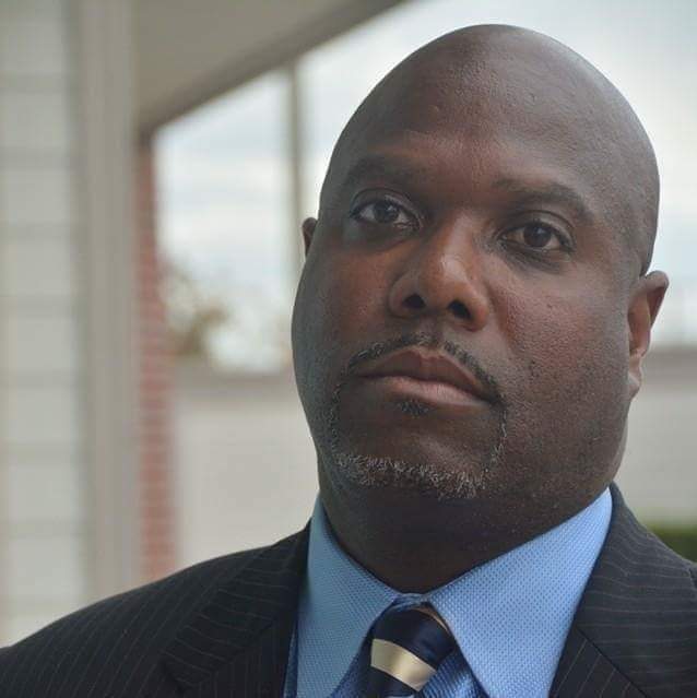A black man wearing a suit and light blue collared shirt looks at the camera