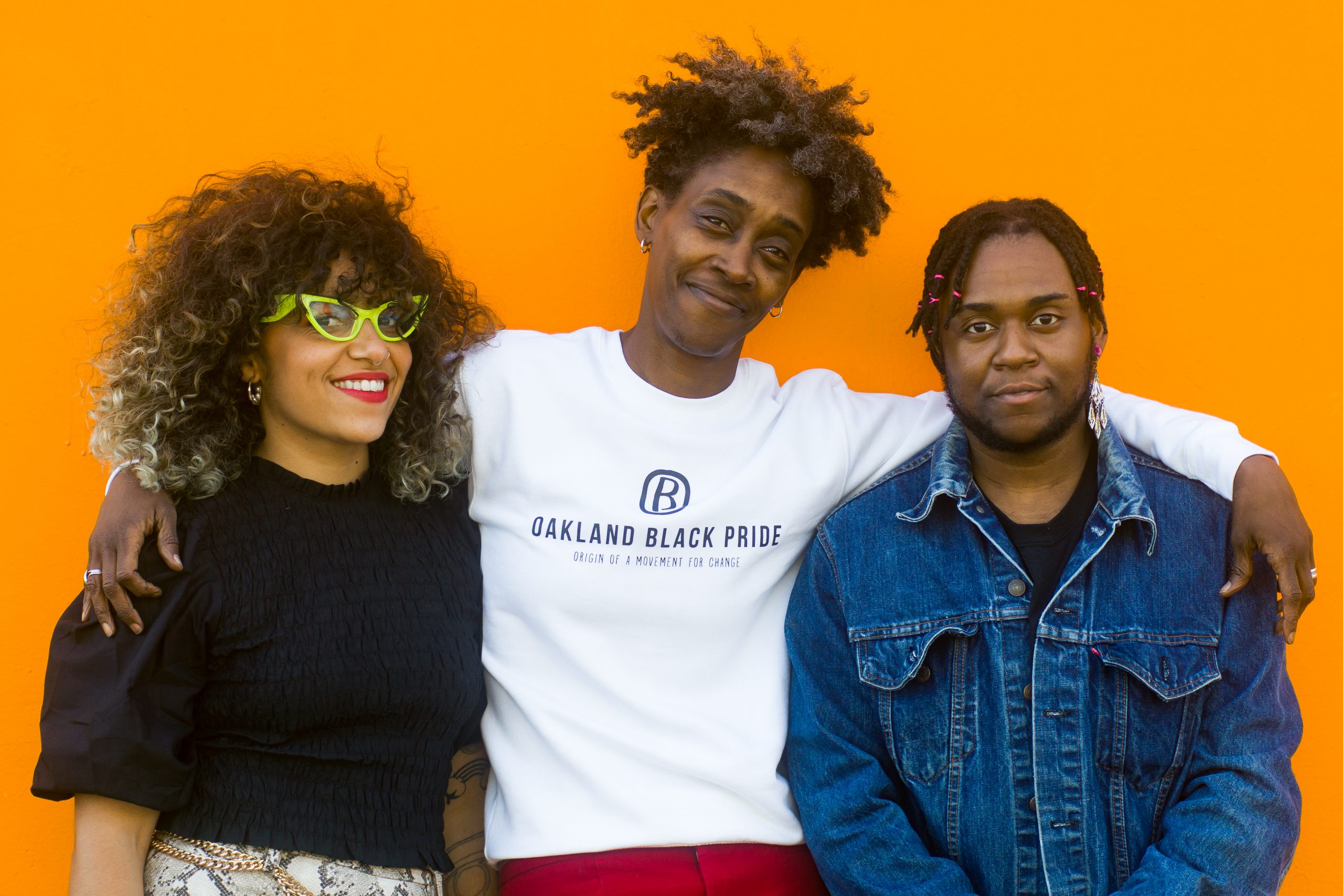 Three Black folks stand against a bright orange background with arms round each other