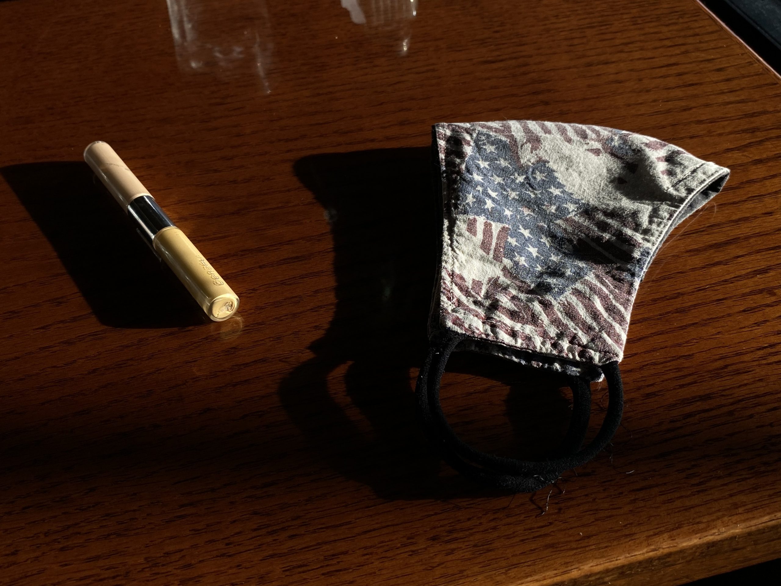 a dark brown wood table has a concealer pen and cloth mask with a faded American flag design