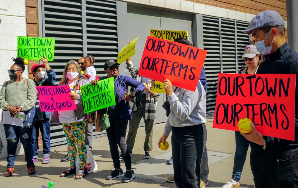 a group of people rally outside a building holding colorful signs with baseball puns asking for benefits to the community