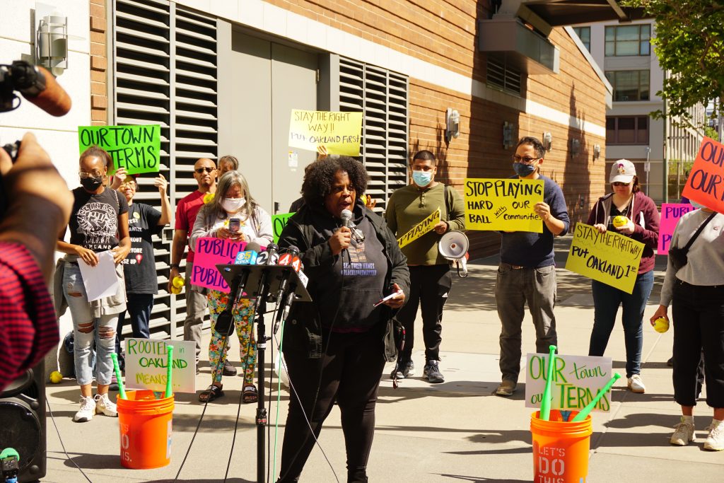 An African American woman speaks into a mic at a rally outside with people behind her holding colorful signs.