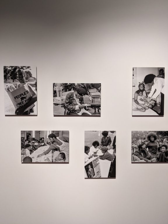 A museum wall with five black and white photos of the Black Panther Party's community programs.