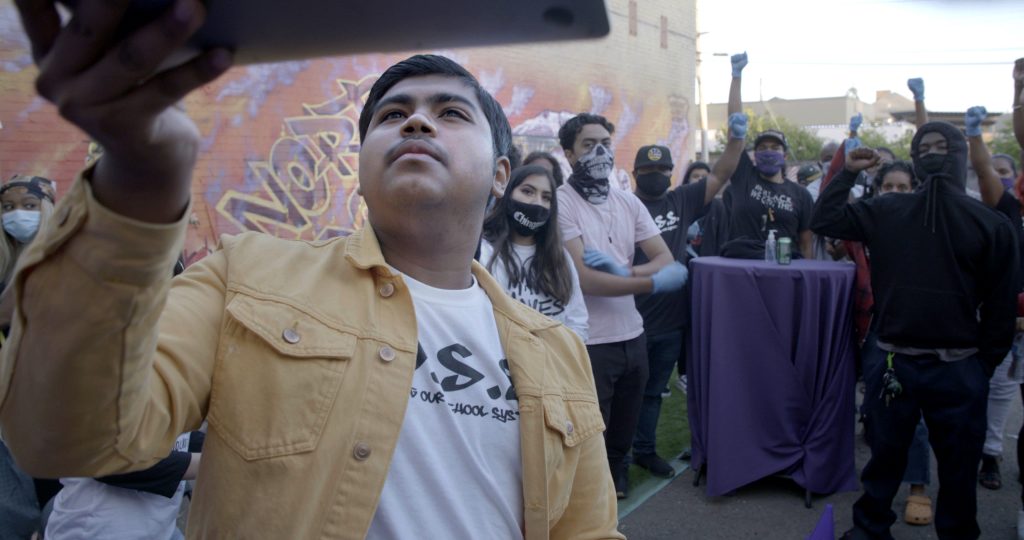 A Latinx youth holds up a screen to take a selfie with a group of people behind him smiling and posing.