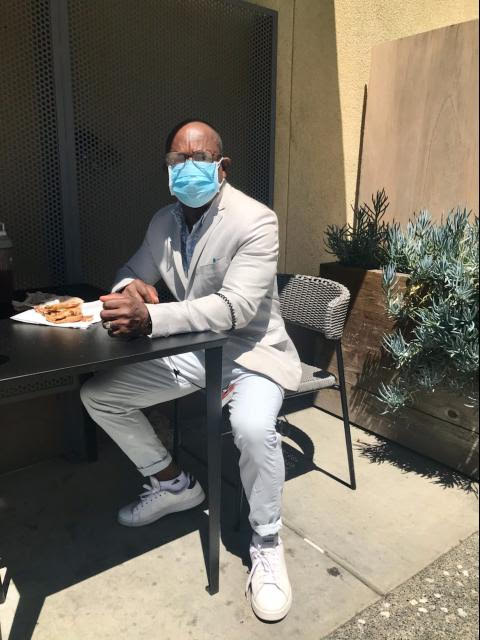 A man sitting in a grey suit coat and pants wearing a mask.