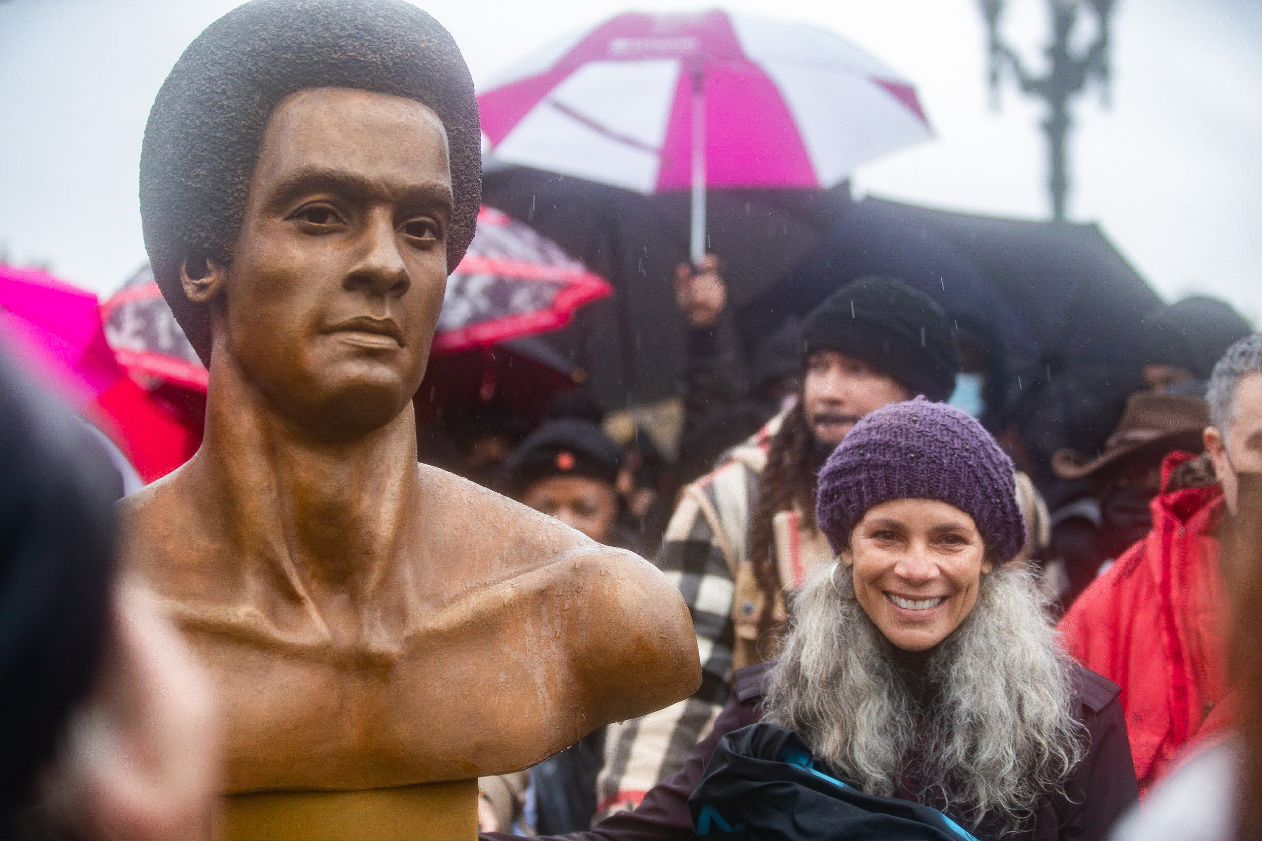 a large shining brown and gold sculpture of a Black man next to petite African American woman who is smiling wearing a purple crocheted hat and flowing curly long grey hair