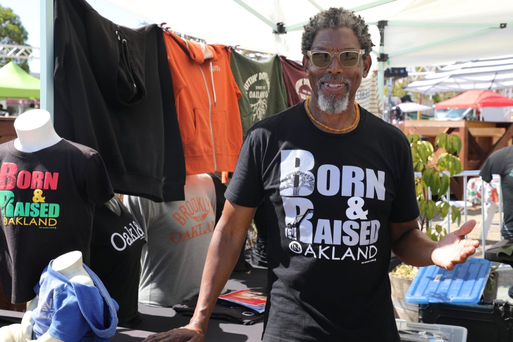 A Black man with short grey hair stands in front of his t-shirt business' stand