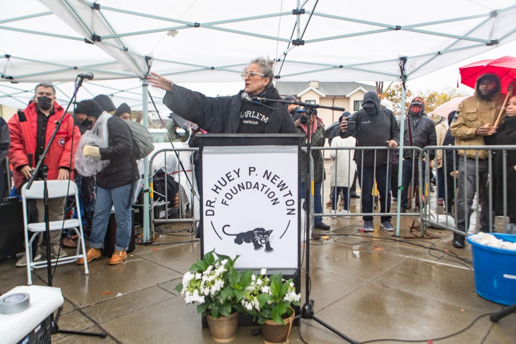A woman with grey hair speaks under a big white tent in the rain with a Black Panther Party banner in front of the podium