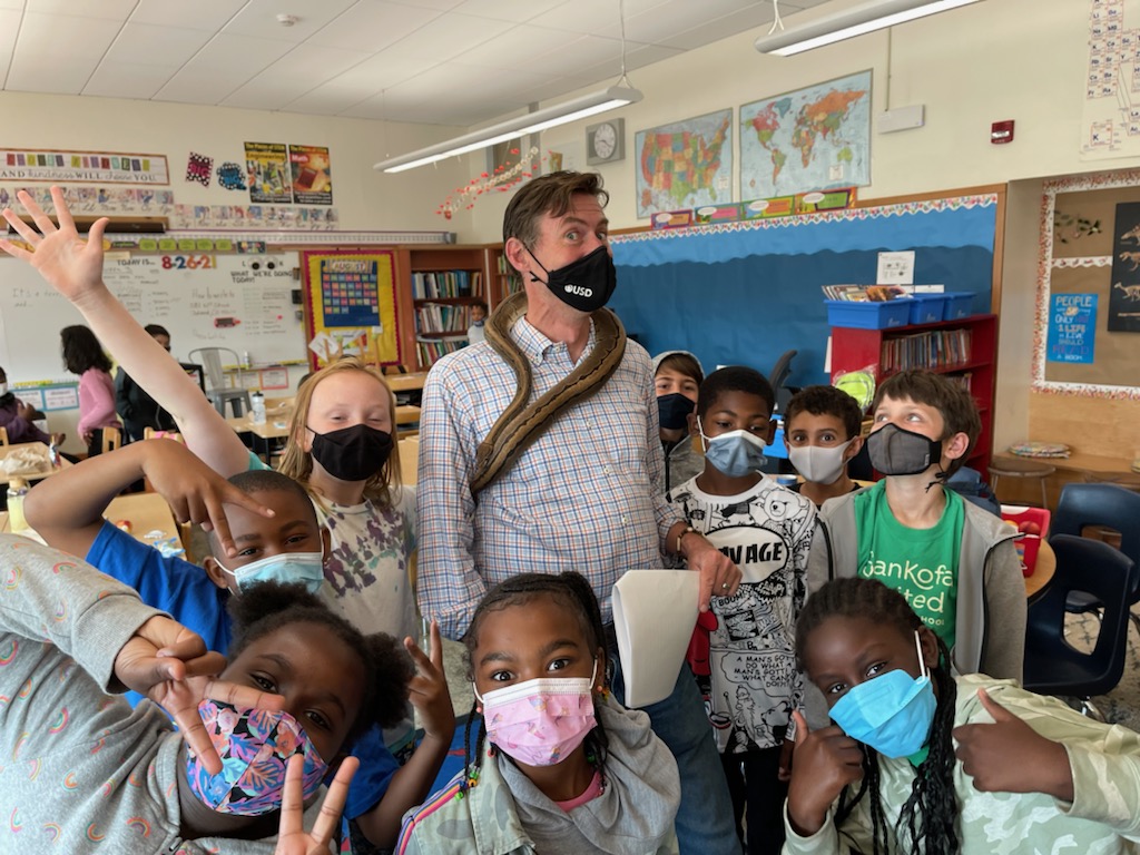 A teacher in a mask is surrounded by happy students in a classroom in Oakland
