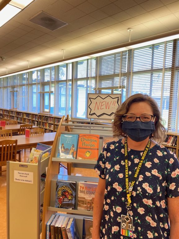 A woman wearing glasses and a mask stands inside a school library.